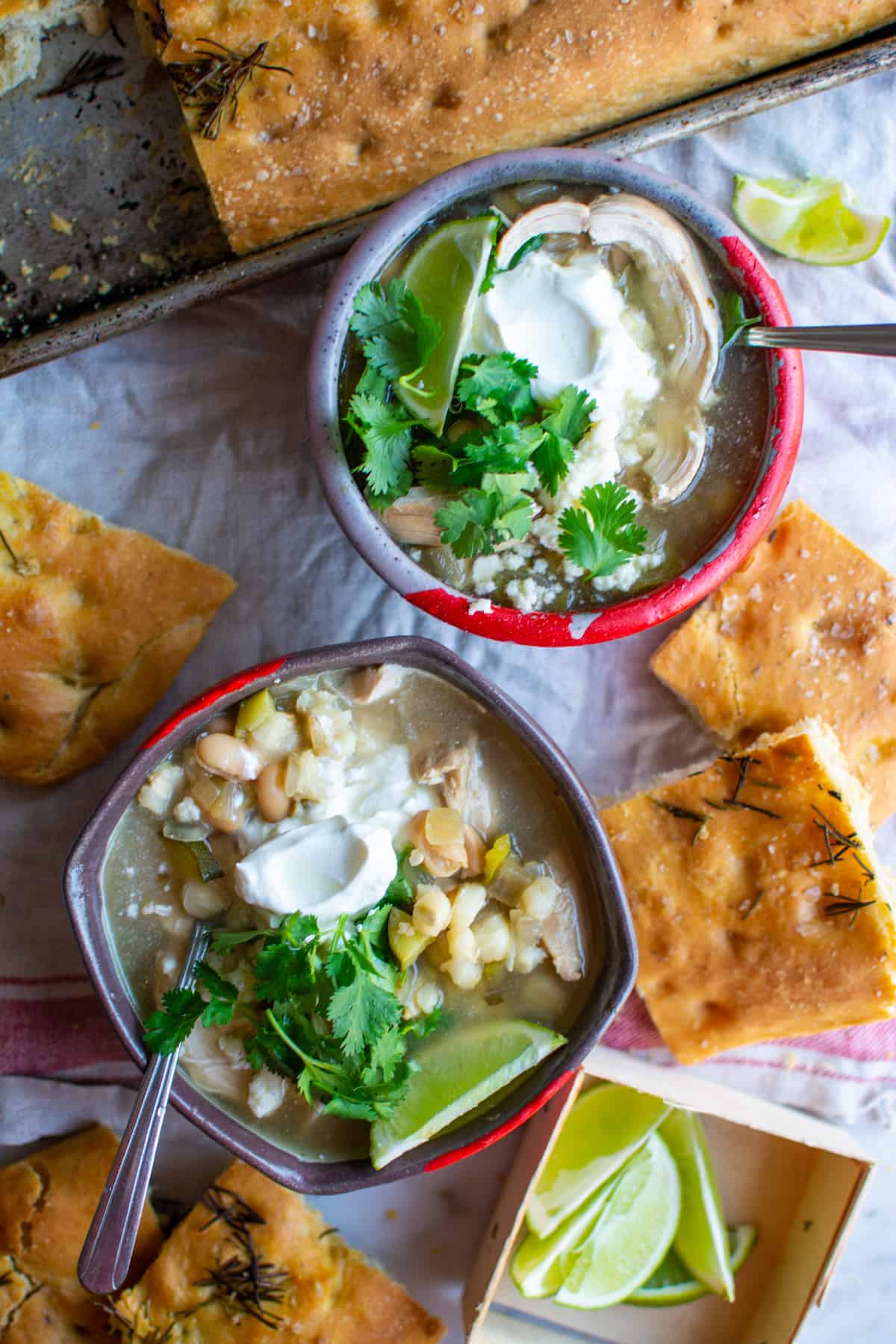 Two bowls of chicken chili on a table covered in a white tablecloth with several pieces of focaccia around it.