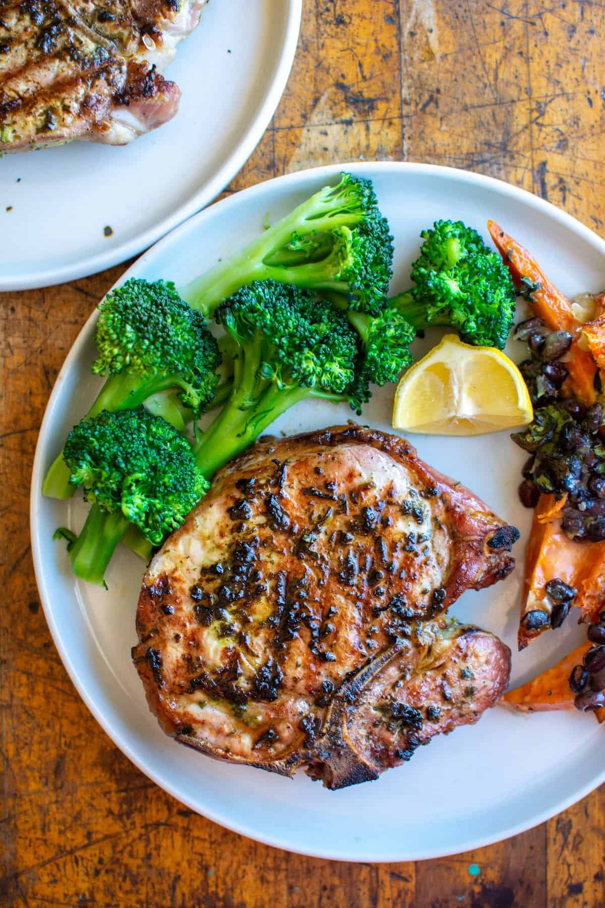 A white plate with a grilled pork chop, broccoli, and a lemon wedge sitting on a wood table with another plate next to it.