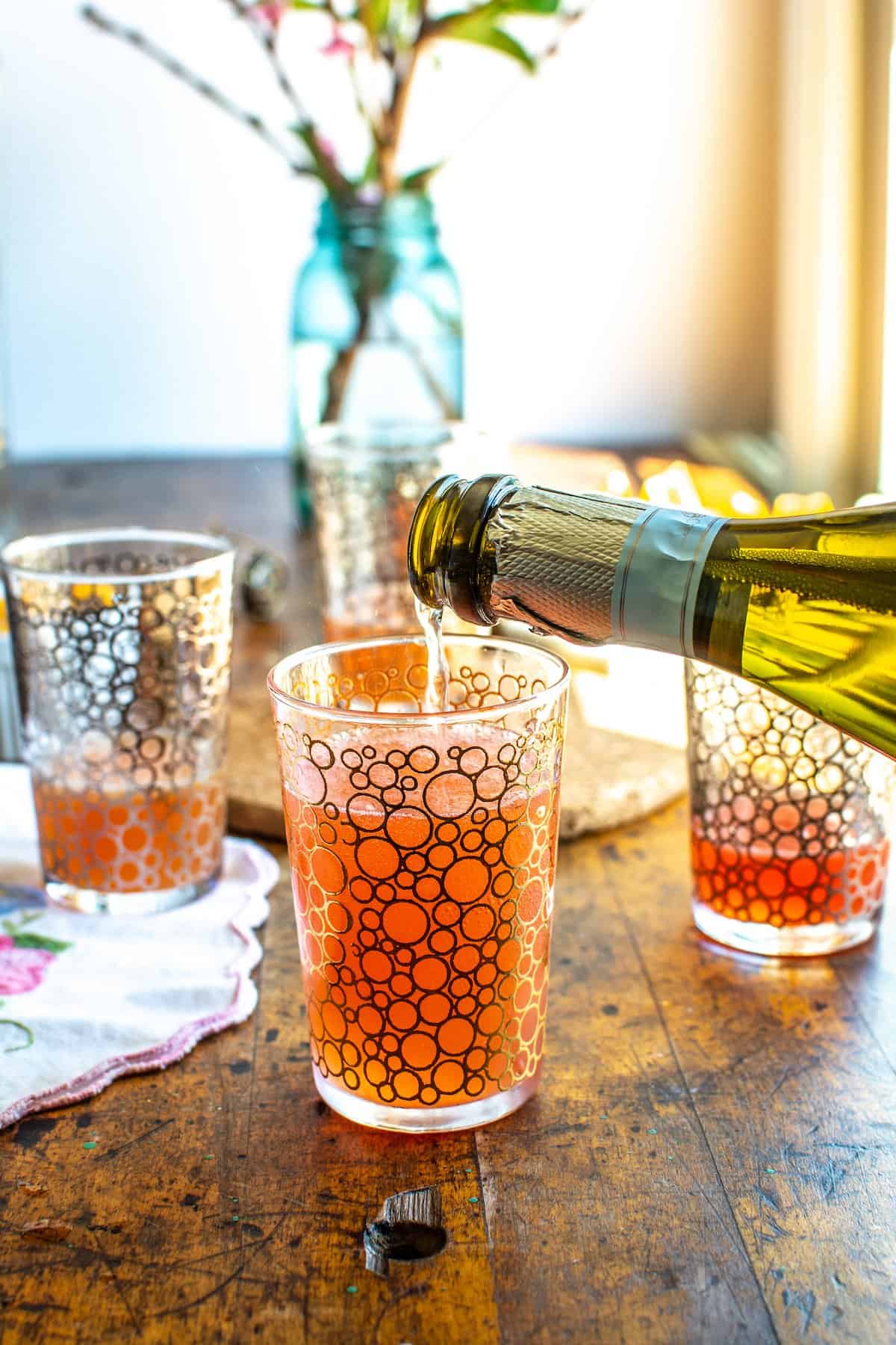 A glass with gold circles filled with a mimosa recipe with champagne being poured into the glass.