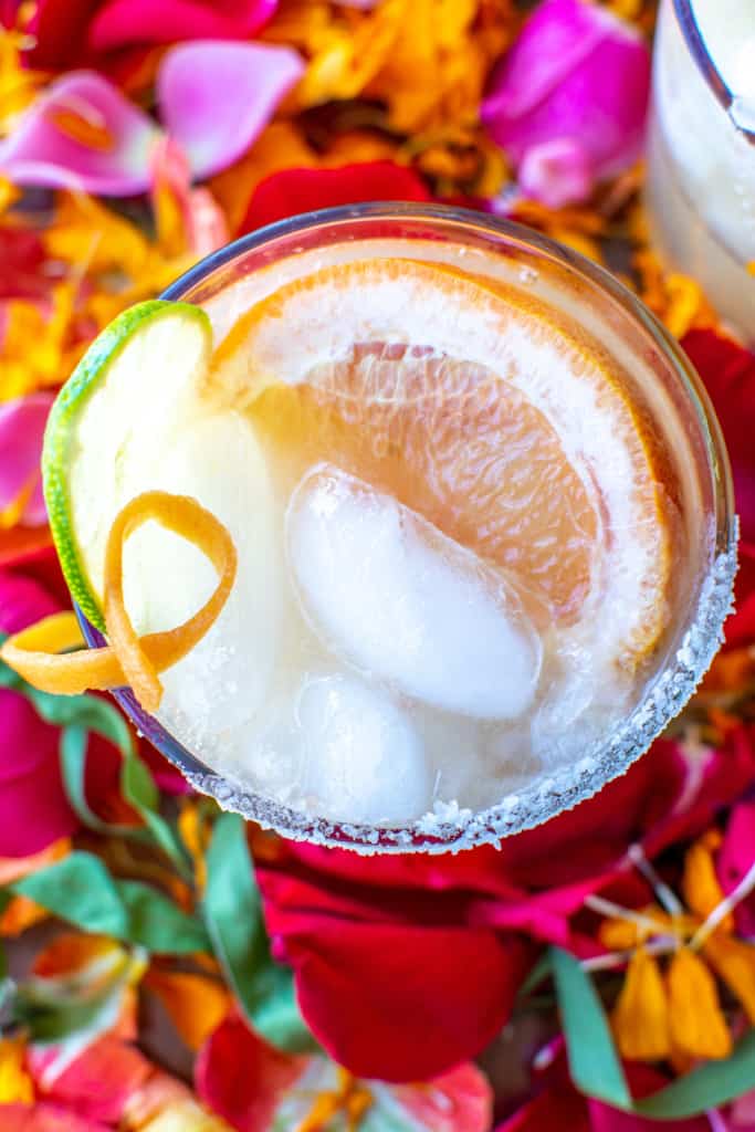 A Honey Grapefruit margarita sitting on a bed of flower petals garnished with a grapefruit wedge, a lime wedge, and an orange twist.