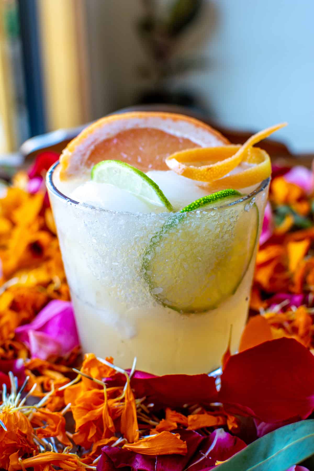 A honey grapefruit margarita sitting on a bed of flower petals with a salt and sugar rimmed glass, a couple slices of lime, and a wedge of grapefruit for garnish.