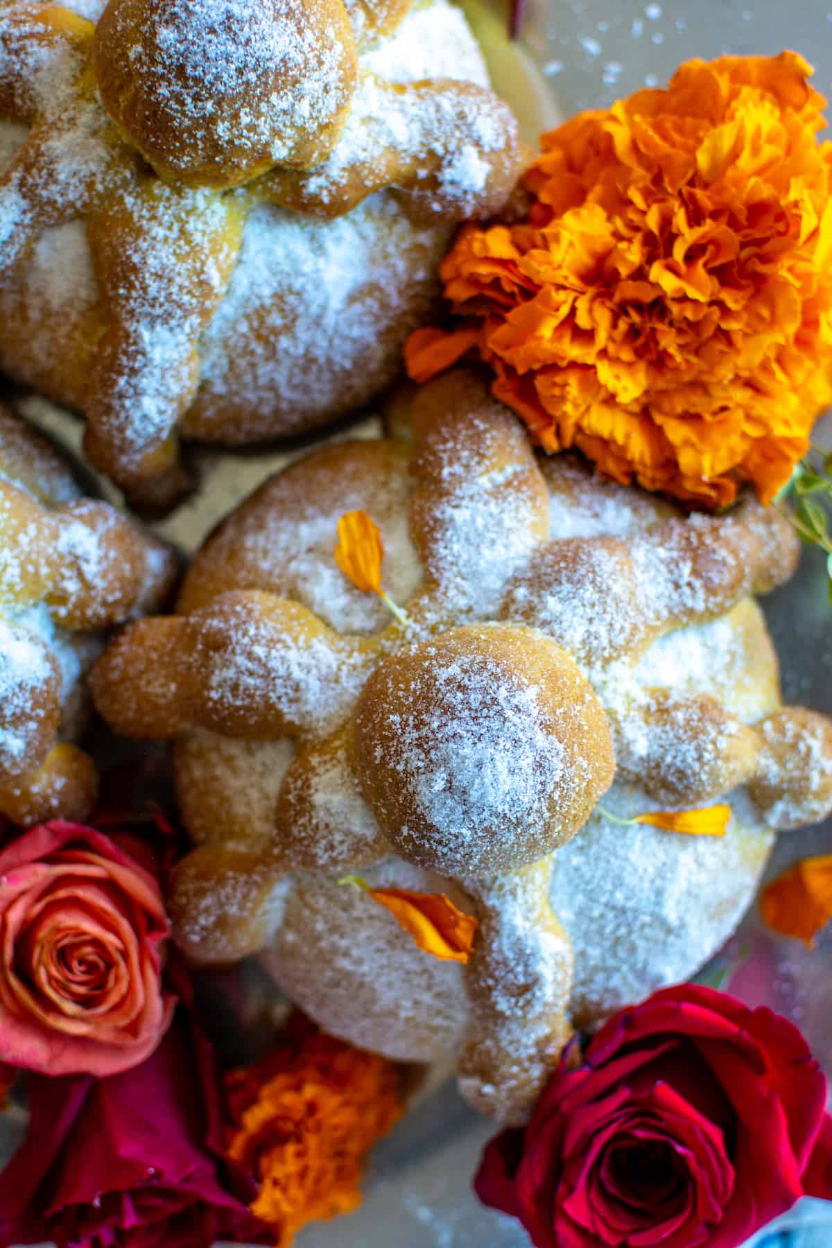 Loaves of pumpkin spice pan de Muerto sitting on a metal tray surrounded by red roses and marigolds.
