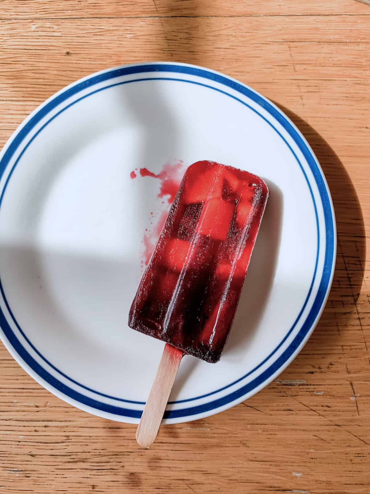 Red popsicle with pieces of mango sitting on a white and blue plate.