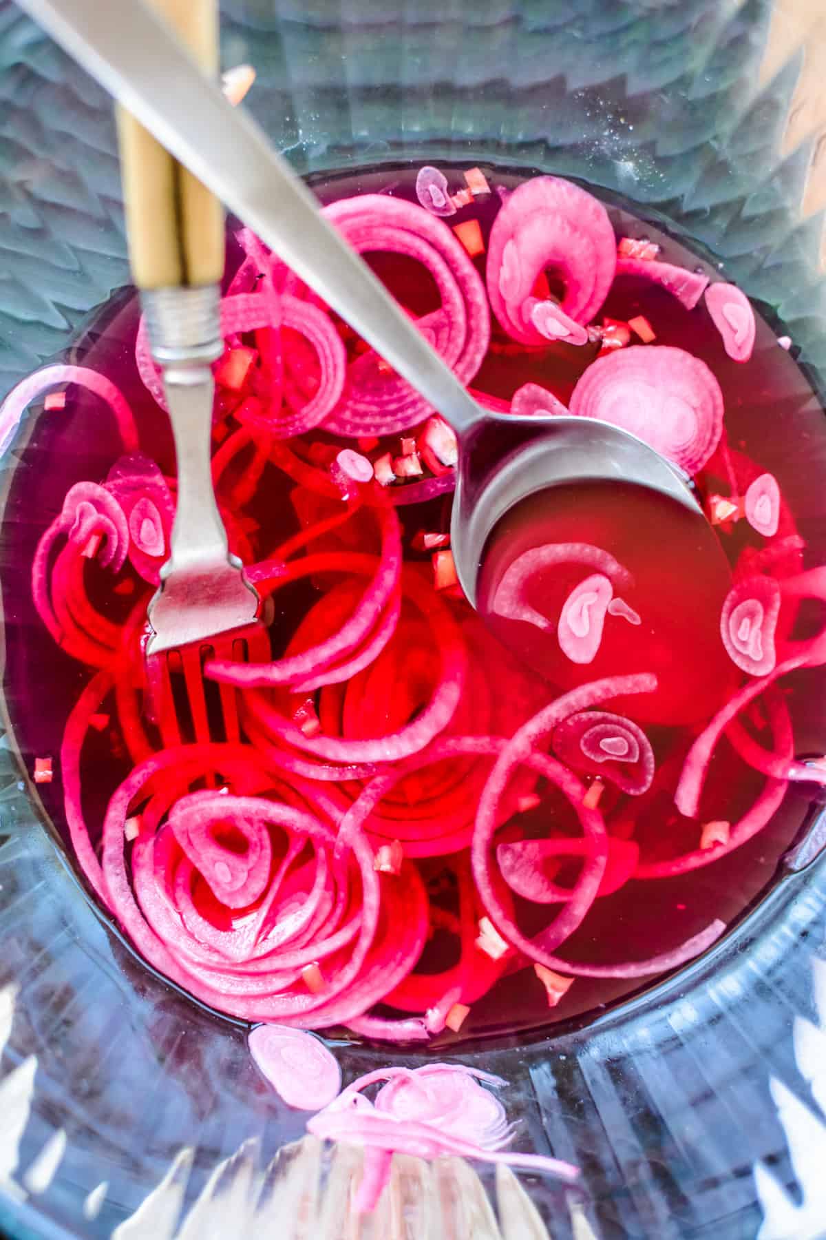 A glass bowl with sliced pickled red onions , a fork, and a spoon inside of it.