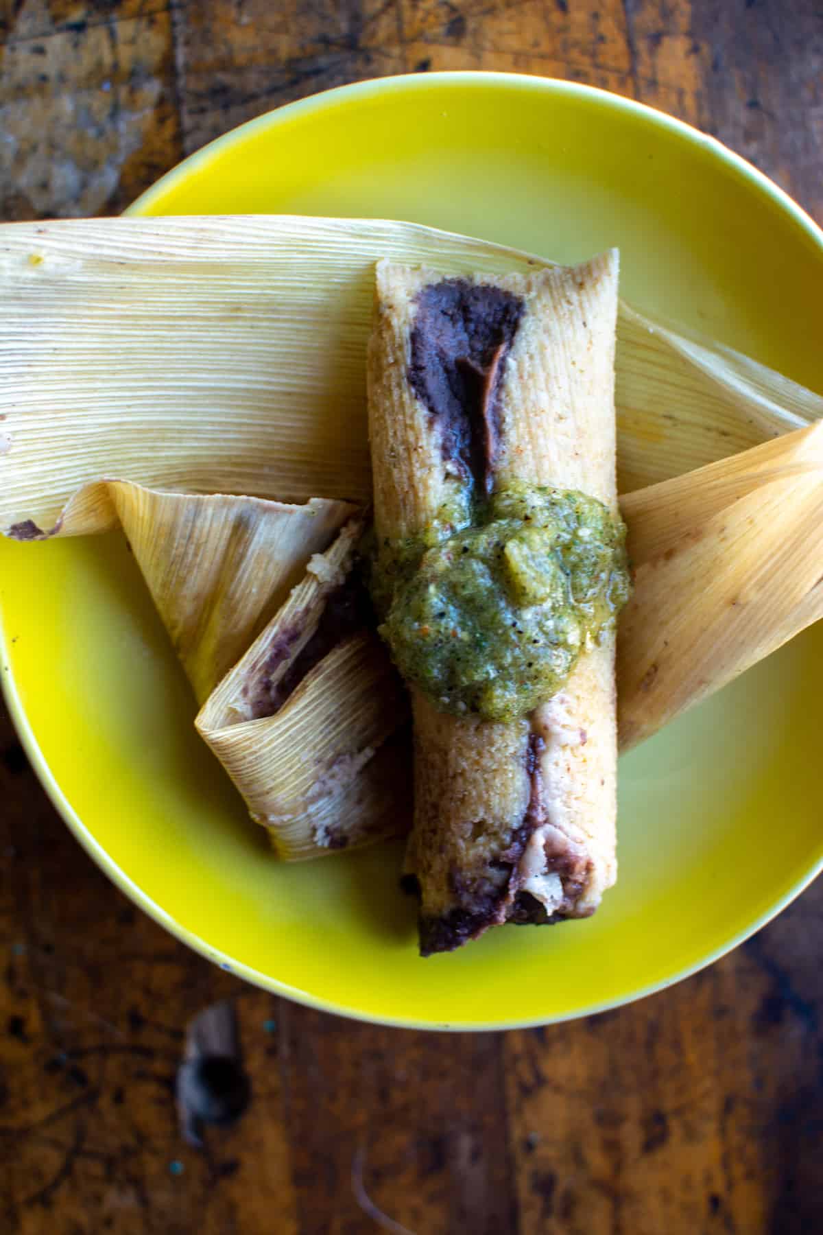 A bean and cheese tamal sitting on top of a corn husk, topped with salsa verde on a yellow plate.