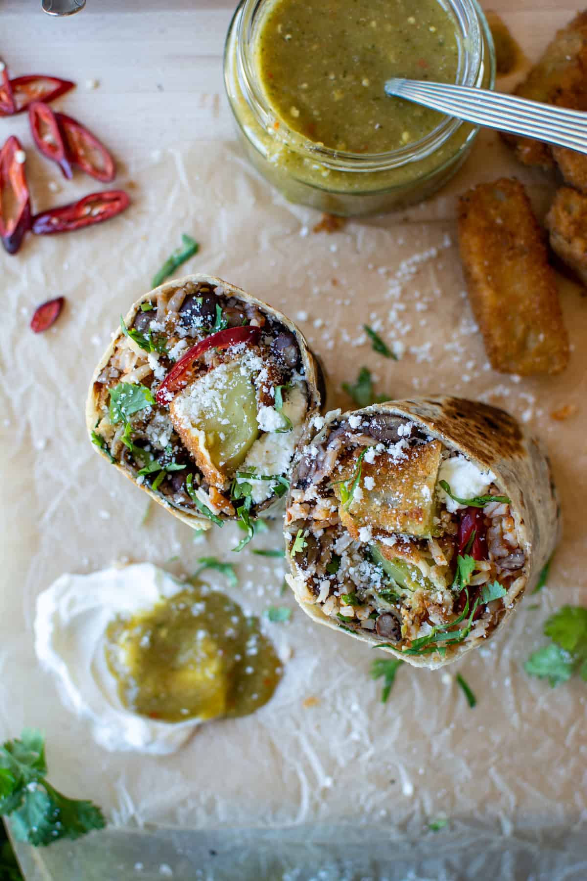 A green tomato burrito cut in half to show the filling on the inside with a jar of salsa on the side and some sliced chiles.