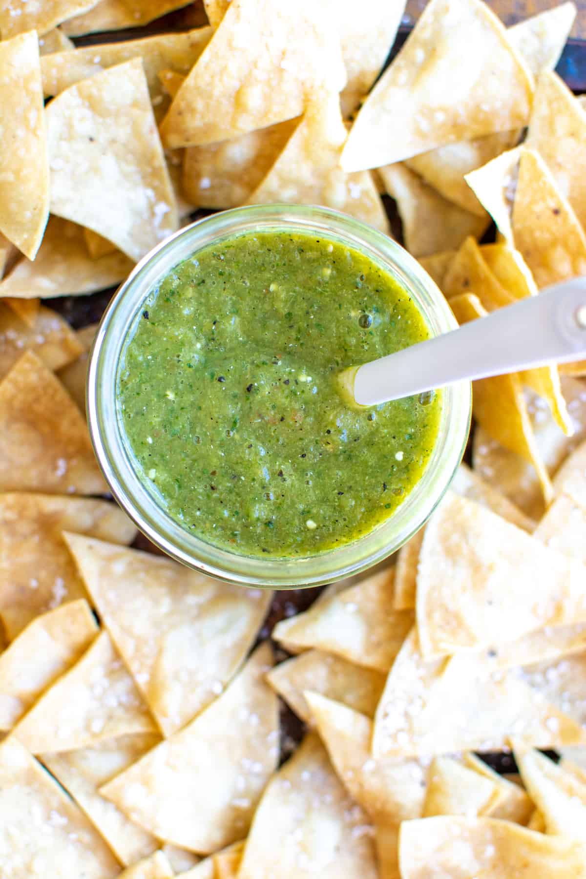 A jar of green salsa sitting in the middle of a large pile of tortilla chips.