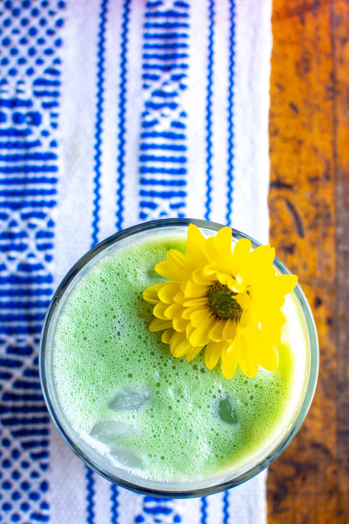 A glass of green Matcha Horchata with a yellow flower in the glass. The glass is sitting on a blue and white runner on a wood table.