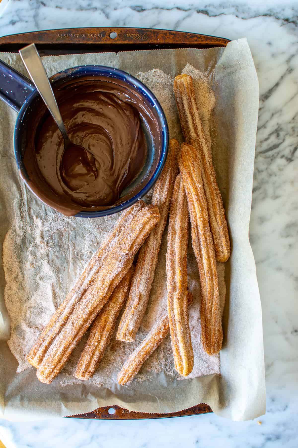 Several churros on a parchment-lined baking sheet with a blue saucepan of chocolate sauce next to them.