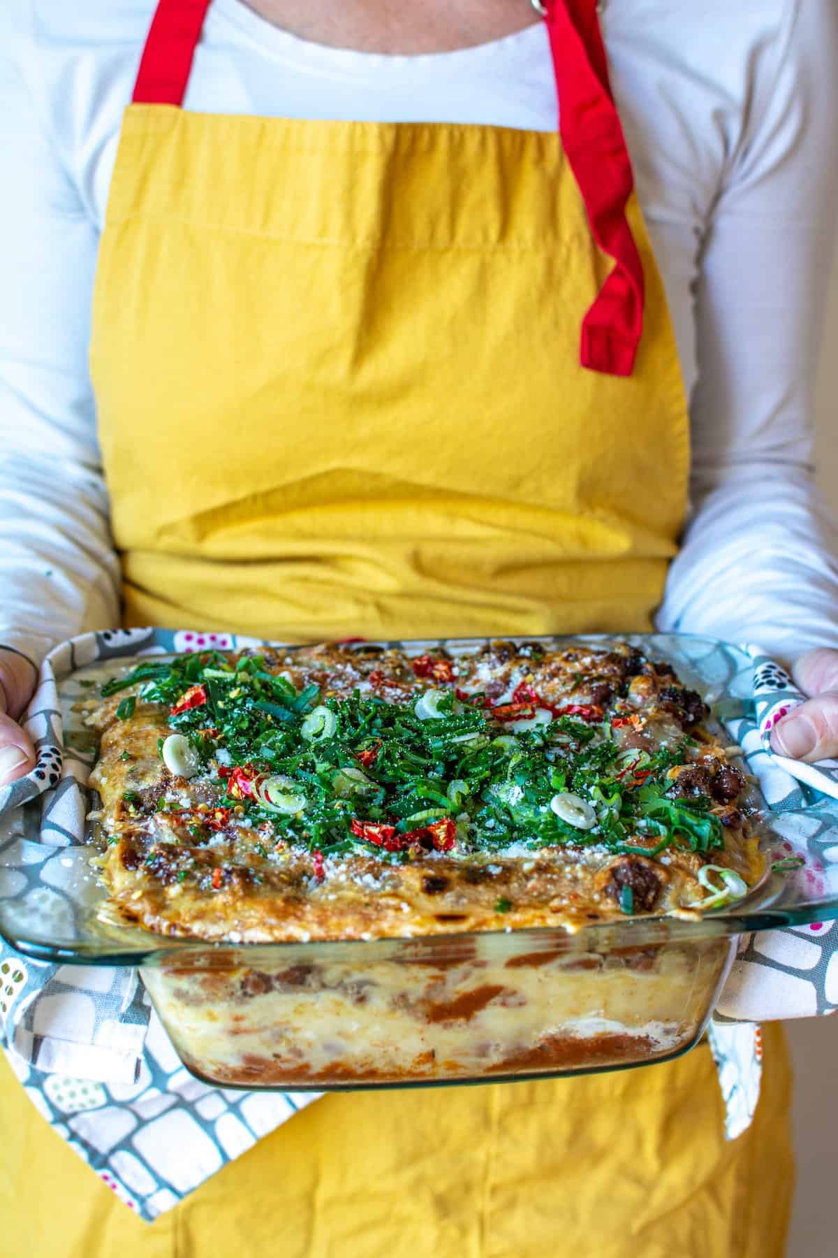 A woman in a yellow apron holding a beef and bean casserole with green onions on top.