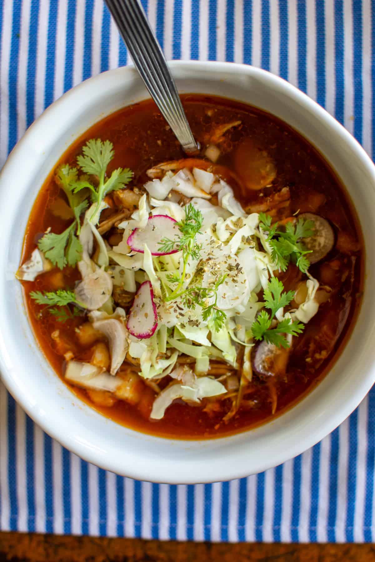 A white bowl of pozole topped with cabbage on a blue and white stripped cloth with a spoon in the bowl.
