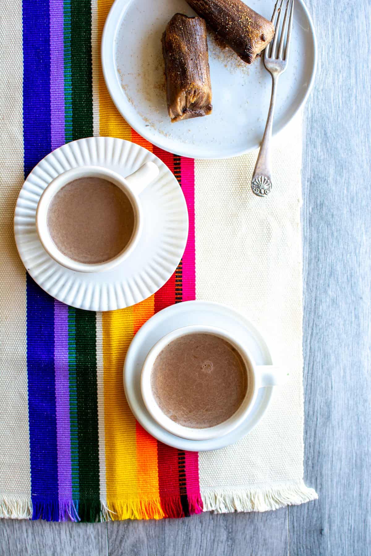 Two white cups of Champurrado sitting on white dishes on a rainbow-striped placemat with a white plate of tamales sitting behind them.