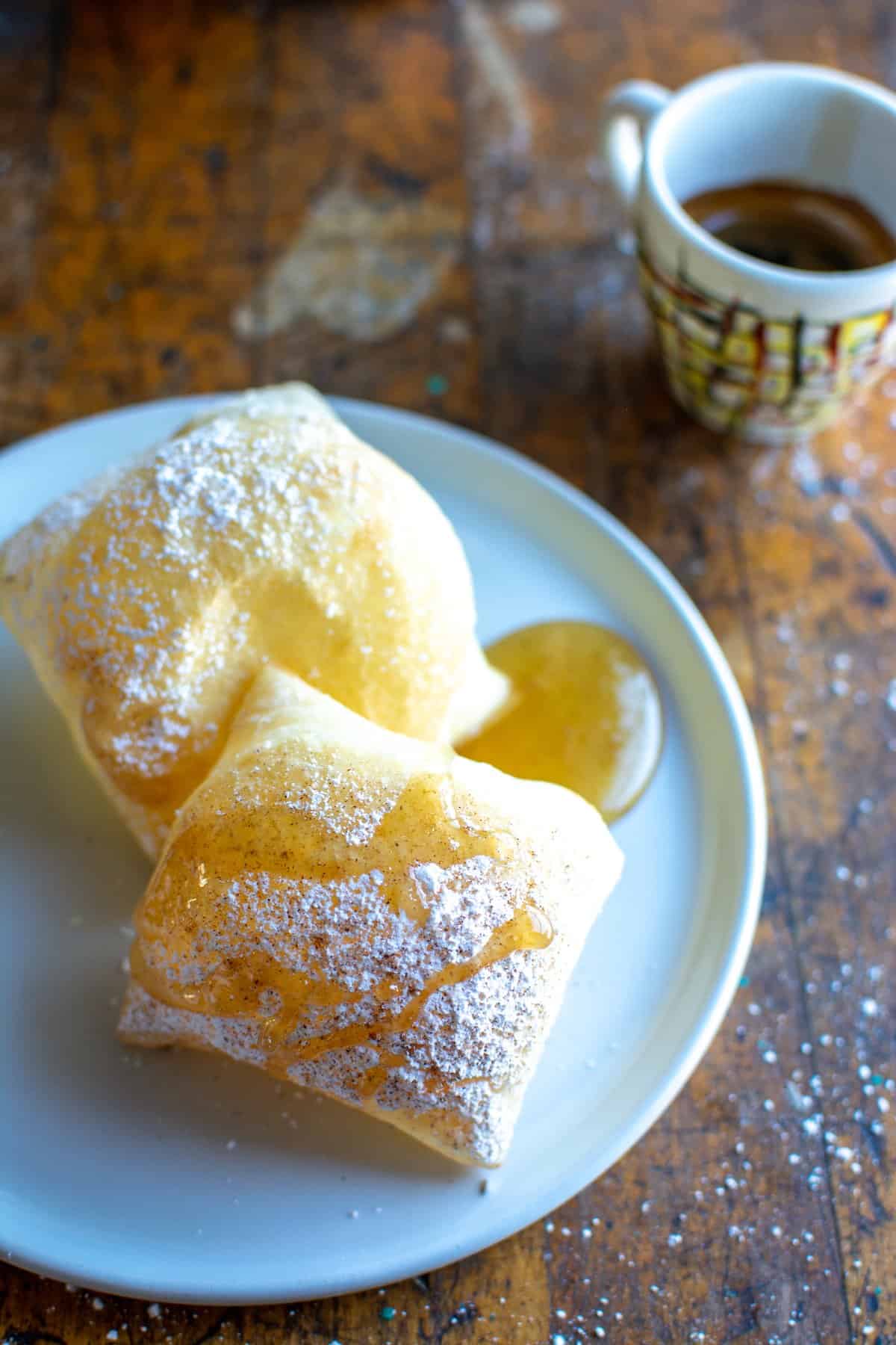Two sopapillas on a white plate drizzled with honey and powdered sugar with a cup of espresso behind it.
