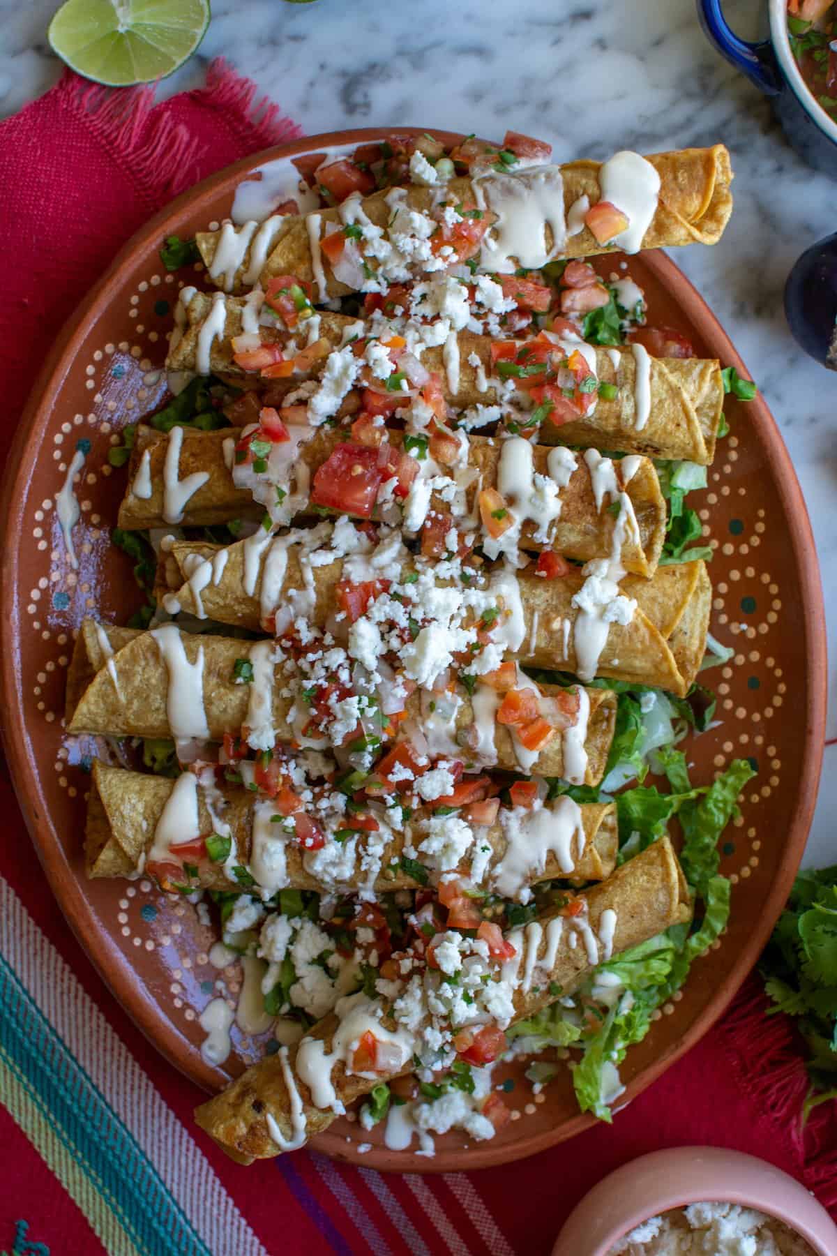 A terracotta plate of flautas Mexicanas topped with sour cream, Pico de Gallo, and queso fresco on a red tablecloth.