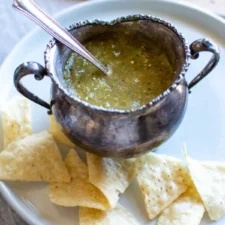 A silver dish filled with habanero salsa on a white plate with some tortilla chips next to it.
