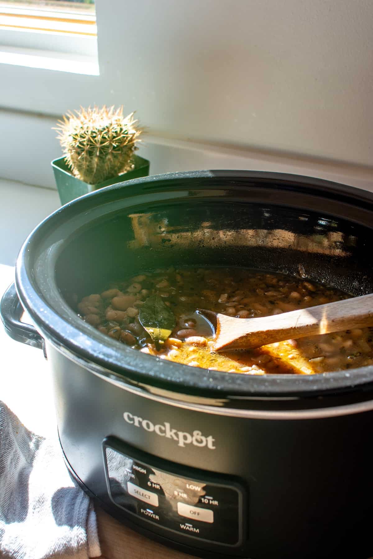 A crockpot full of beans sitting on the counter with a wood spoon in it.