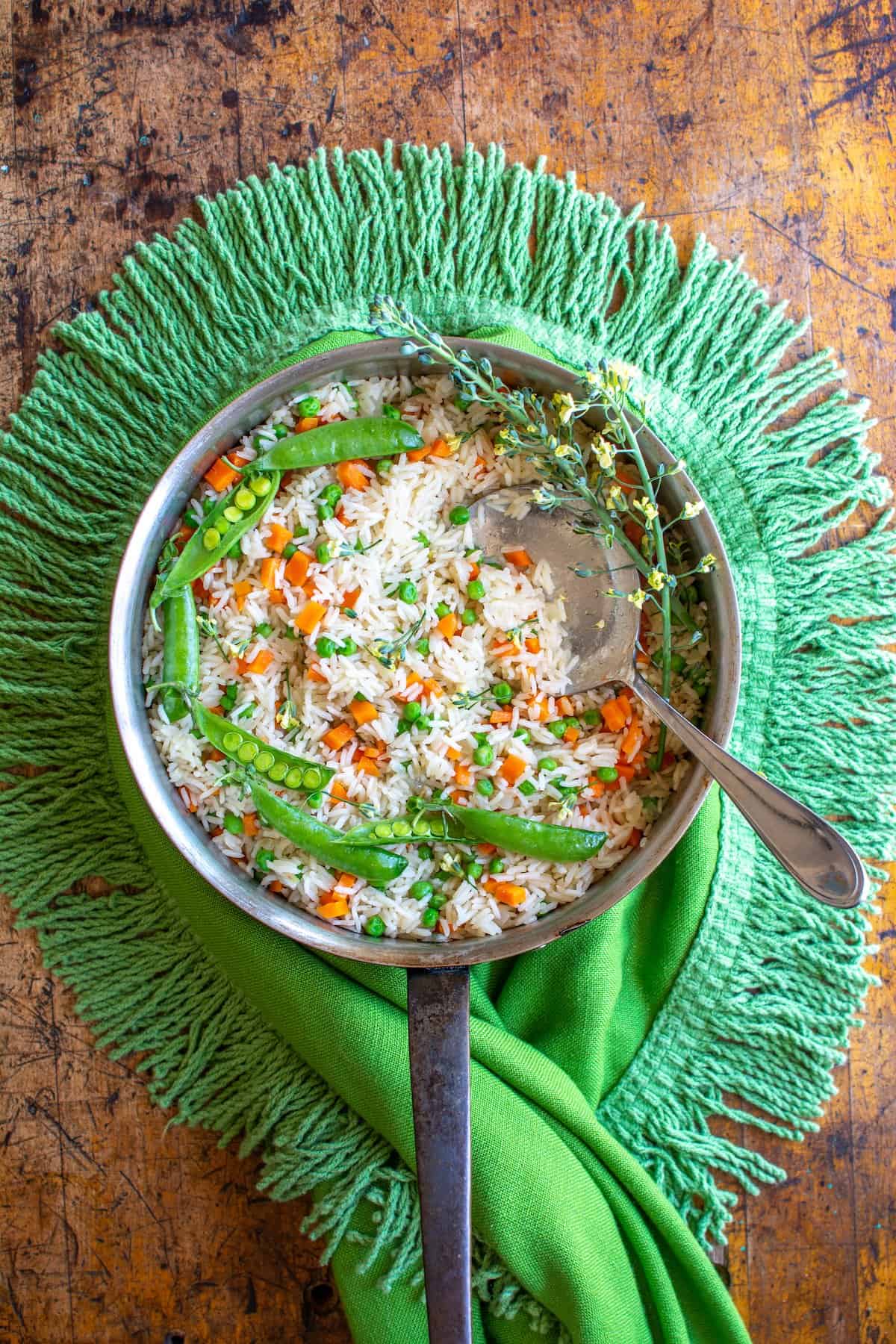 A pot of Arroz with carrots and peas sitting on a green tablecloth on a wood table with a spoon sticking out of one side.