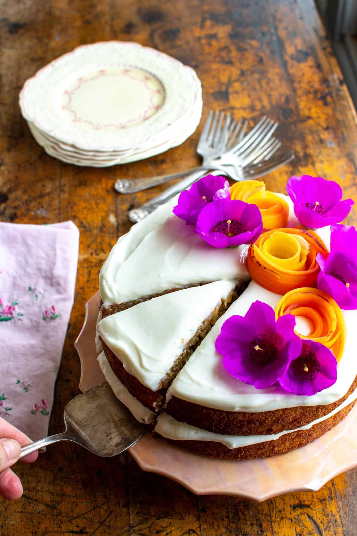 A woman removing a piece of cake from a carrot cake topped with spirals of carrots and purple flowers.