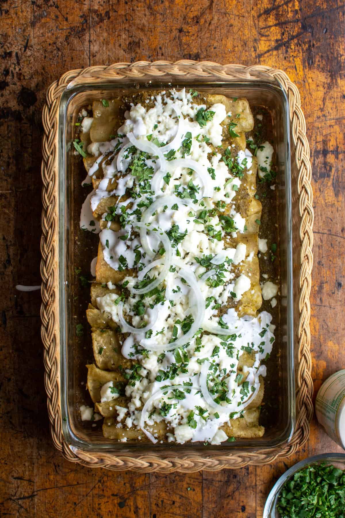A baking dish filled with enchiladas verdes covered in a whicker basket on a wood table.