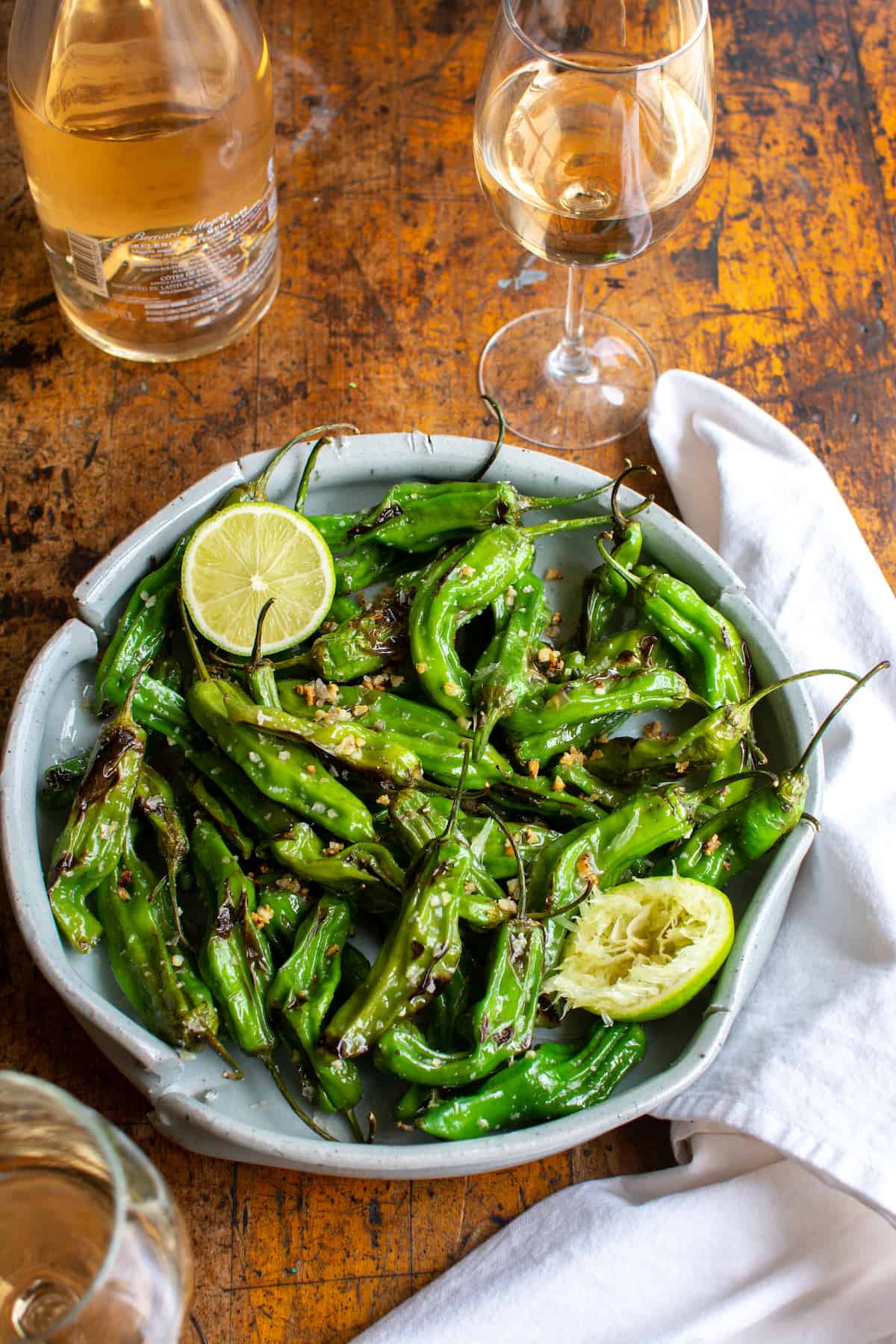 A large platter of shishito peppers with a lime cut in half on the platter next to a glass and bottle of wine.