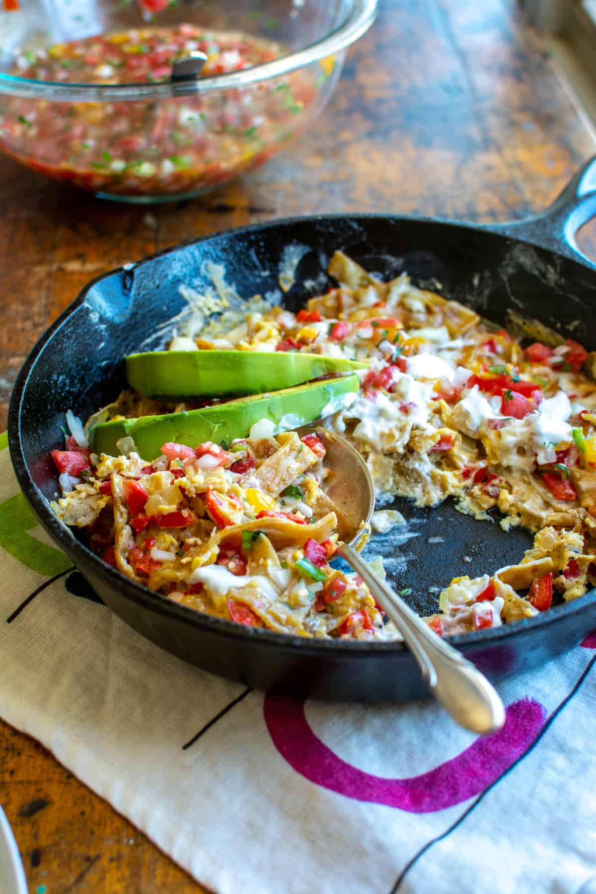 A black cast iron skillet filled with migas on a white kitchen towel with a glass bowl behind it.