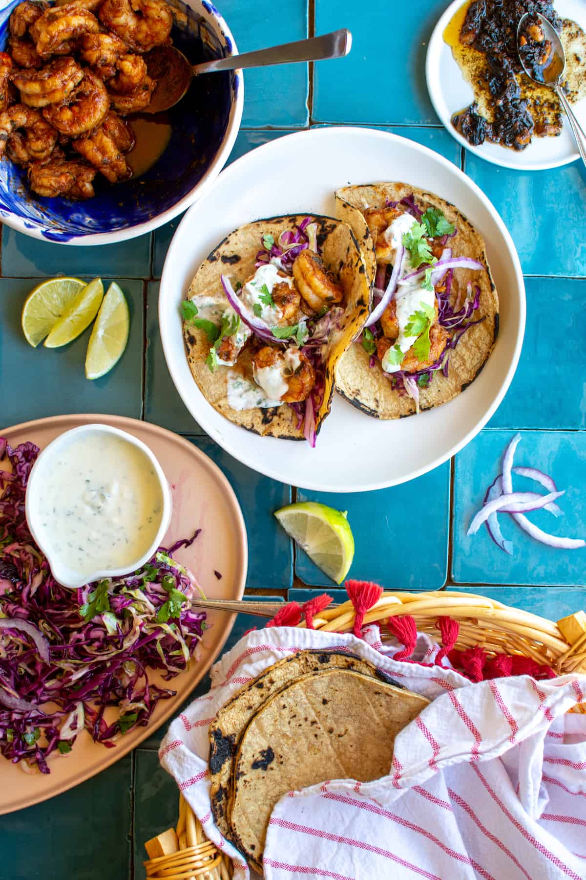 A white plate of shrimp tacos sitting on a blue tiled table next to a bowl of the charred shrimp, a basket of corn tortillas, and some purple cabbage slaw.