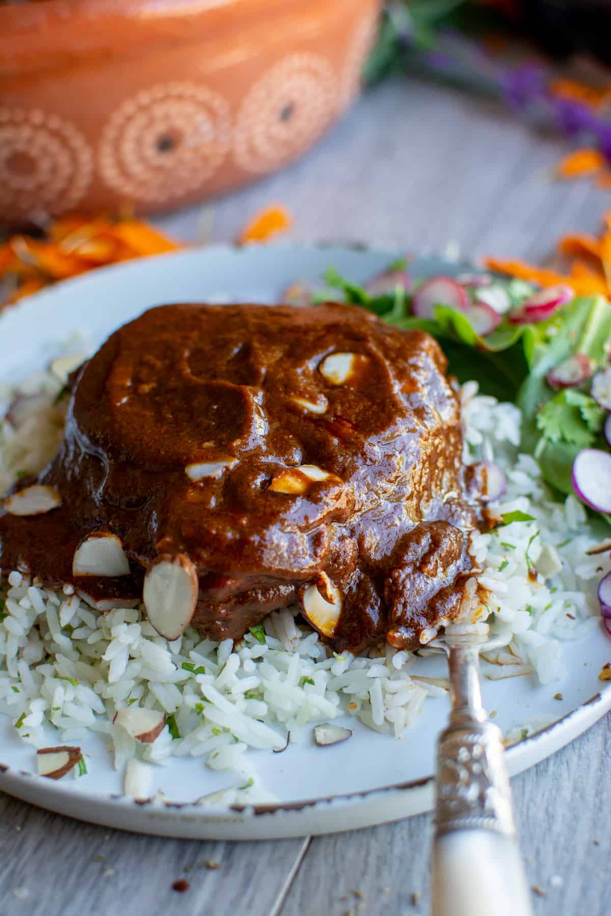 A piece of chicken mole sitting on a bed of rice next to a green Salad with a fork on the plate.