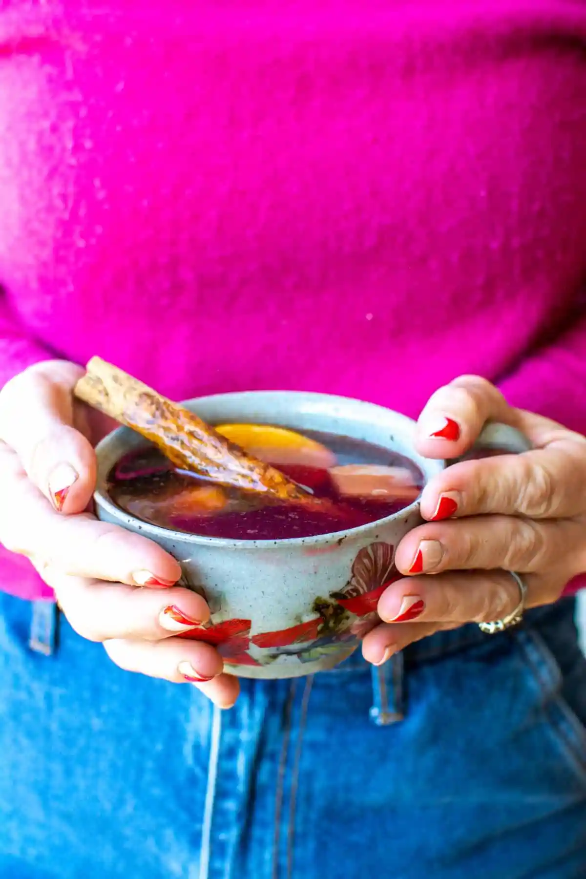A woman in a pink sweater holding a cup of ponche with a cinnamon stick, sticking out of it.