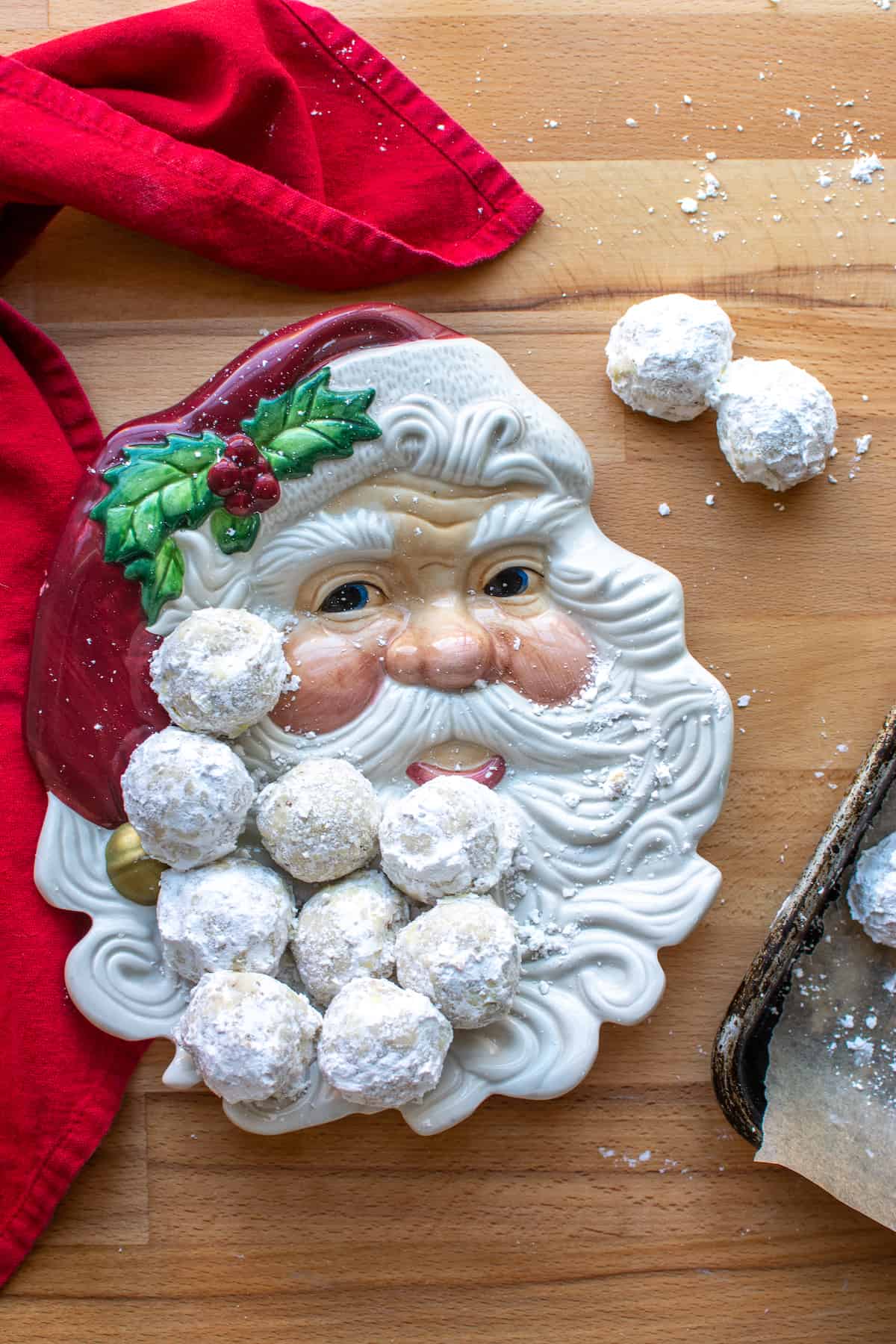 A Santa cookie plate with several Mexican wedding cookies on it sitting on a wood table by a red napkin.