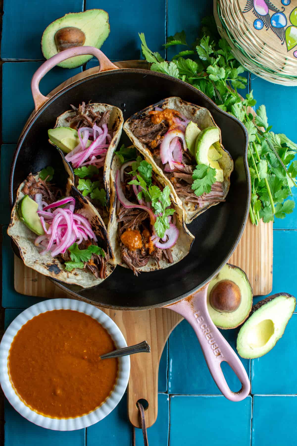 A pink frying pan with four barbacoa tacos in it on a blue tile background