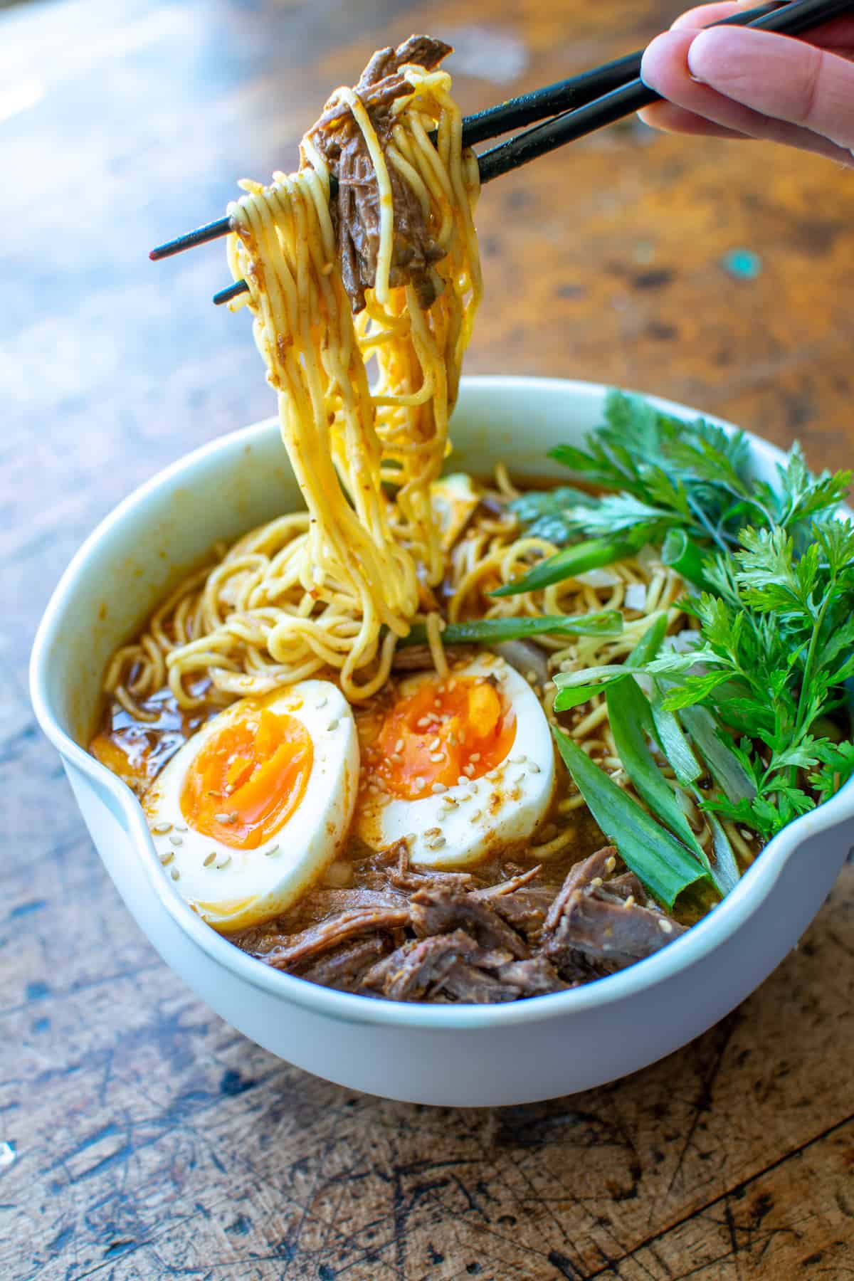 A bowl of Birria Ramen with a person pulling the noodles out with chopsicks.