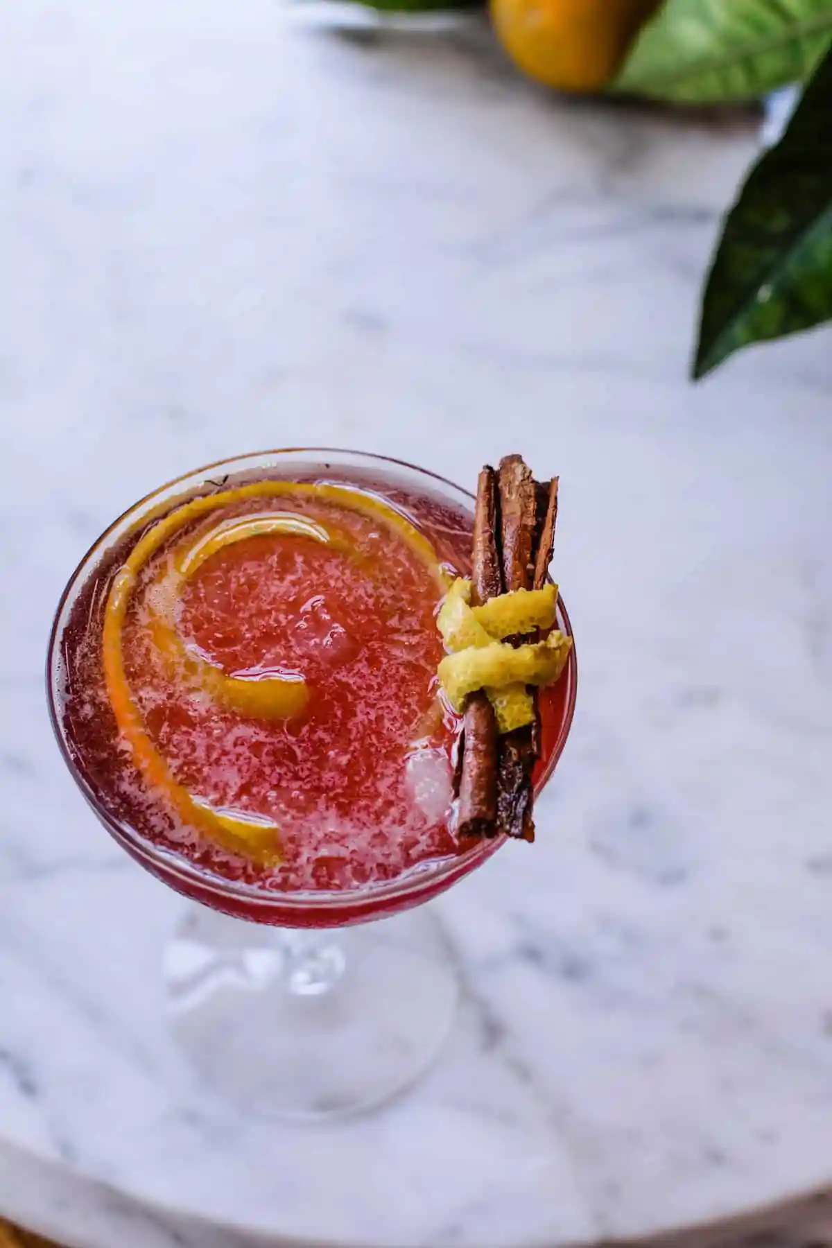 A red cocktail sitting on a marble table with a cinnamon stick wrapped in orange peel.