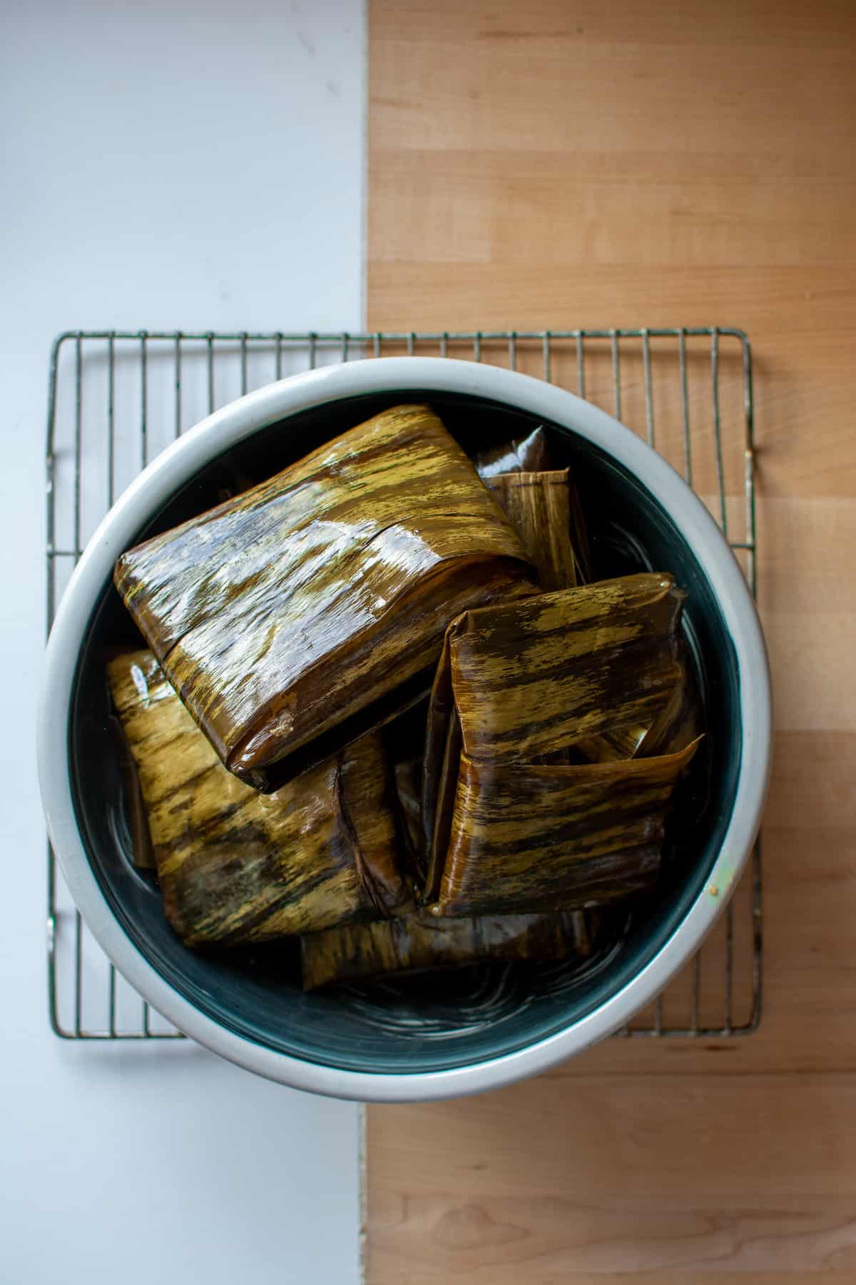 A bowl sitting on a wire rack filled with tamales wrapped in banana leaves.