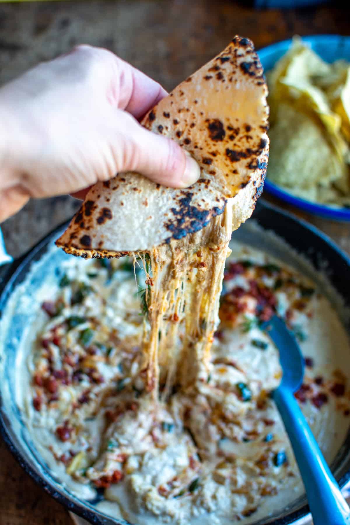 A hand holding a tortilla over a pan of choriqueso with the cheese dripping back into the pan.