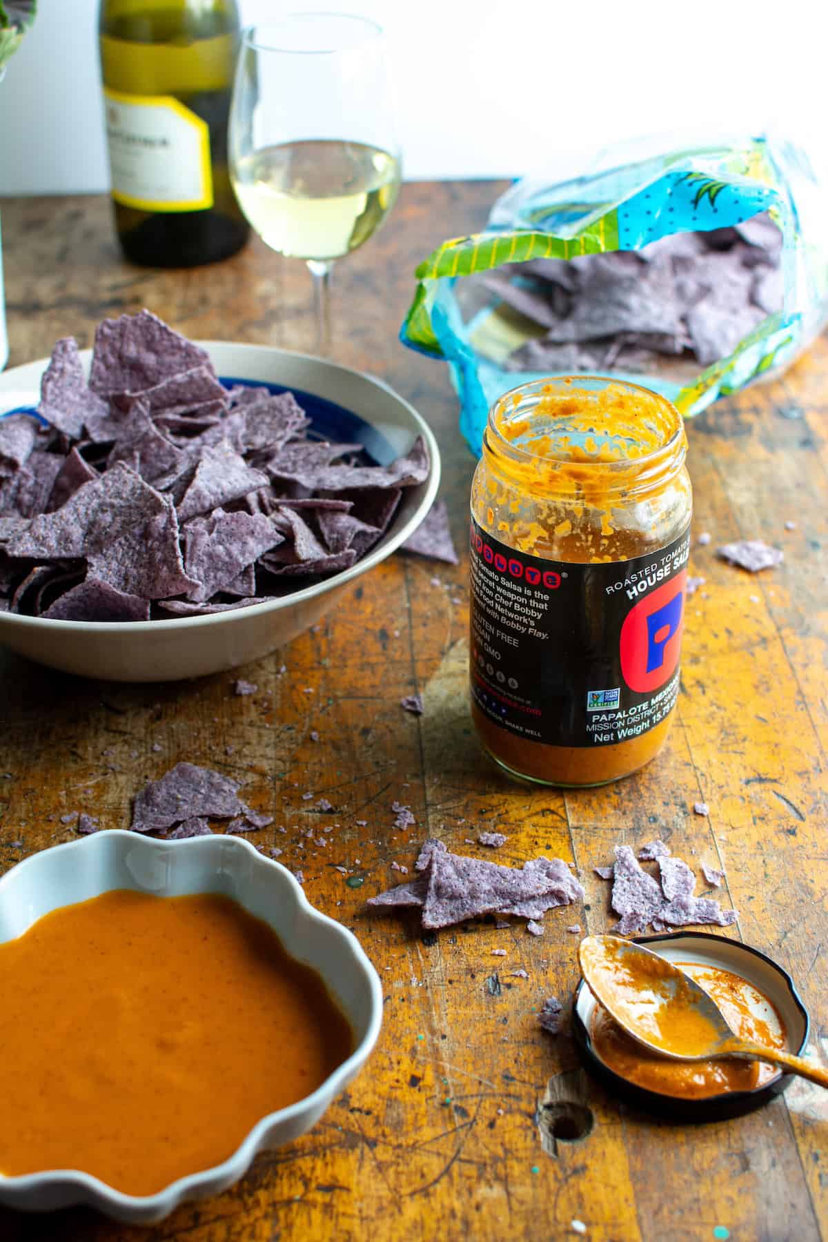 A jar of salsa on a wood table next to a bowl of blue tortilla chips