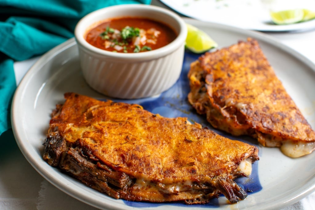 two birria tacos with a bowl of consommé 