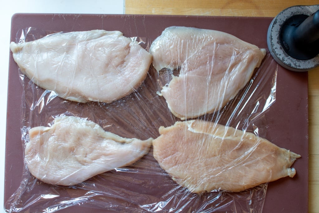chicken breasts on a cutting board covered in plastic wrap